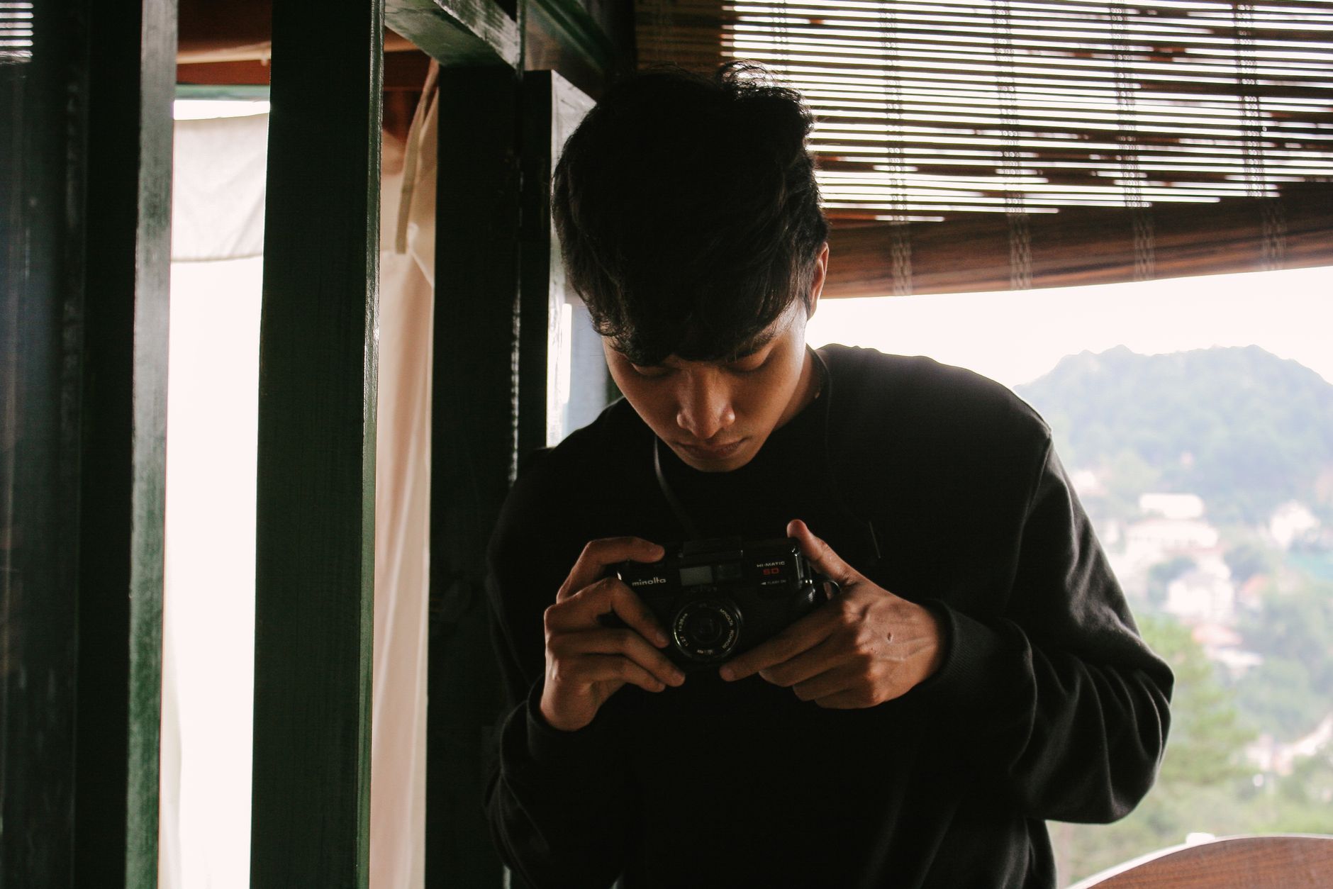 A young man focuses intently on a vintage camera by a window indoors.