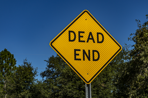 Free Close-up of a yellow 'Dead End' sign against a blue sky and trees, symbolizing caution and warning. Stock Photo