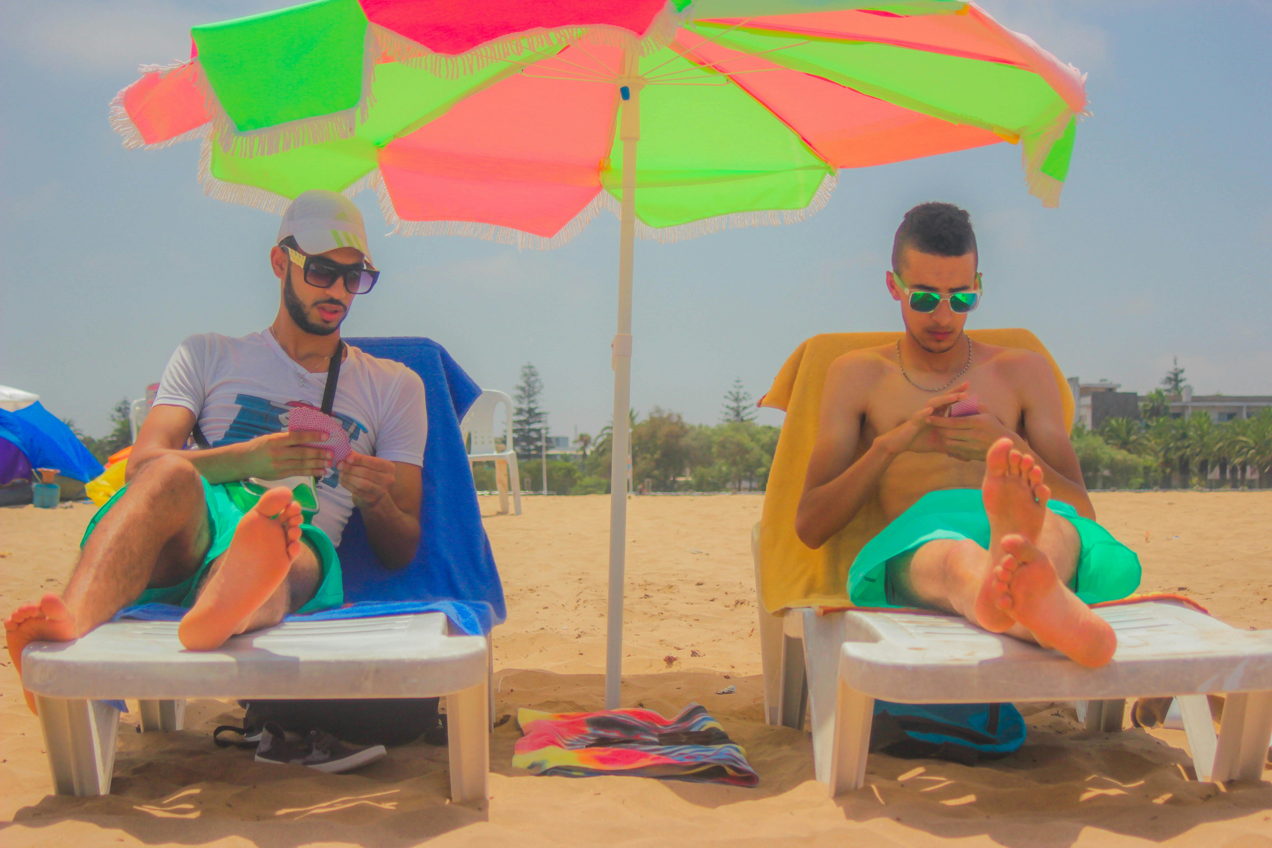 Two young men sitting on beach loungers under a colorful umbrella, enjoying a sunny day.
