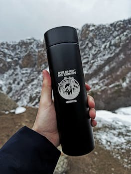Close-up of a hand holding a black thermos against a snowy mountain backdrop.