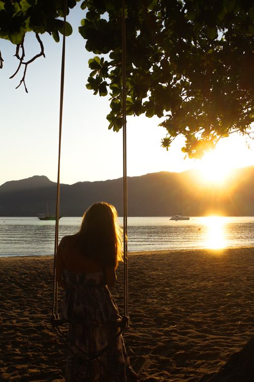 Free A woman sits on a swing by the beach, taking in the serene sunset over the ocean. Stock Photo