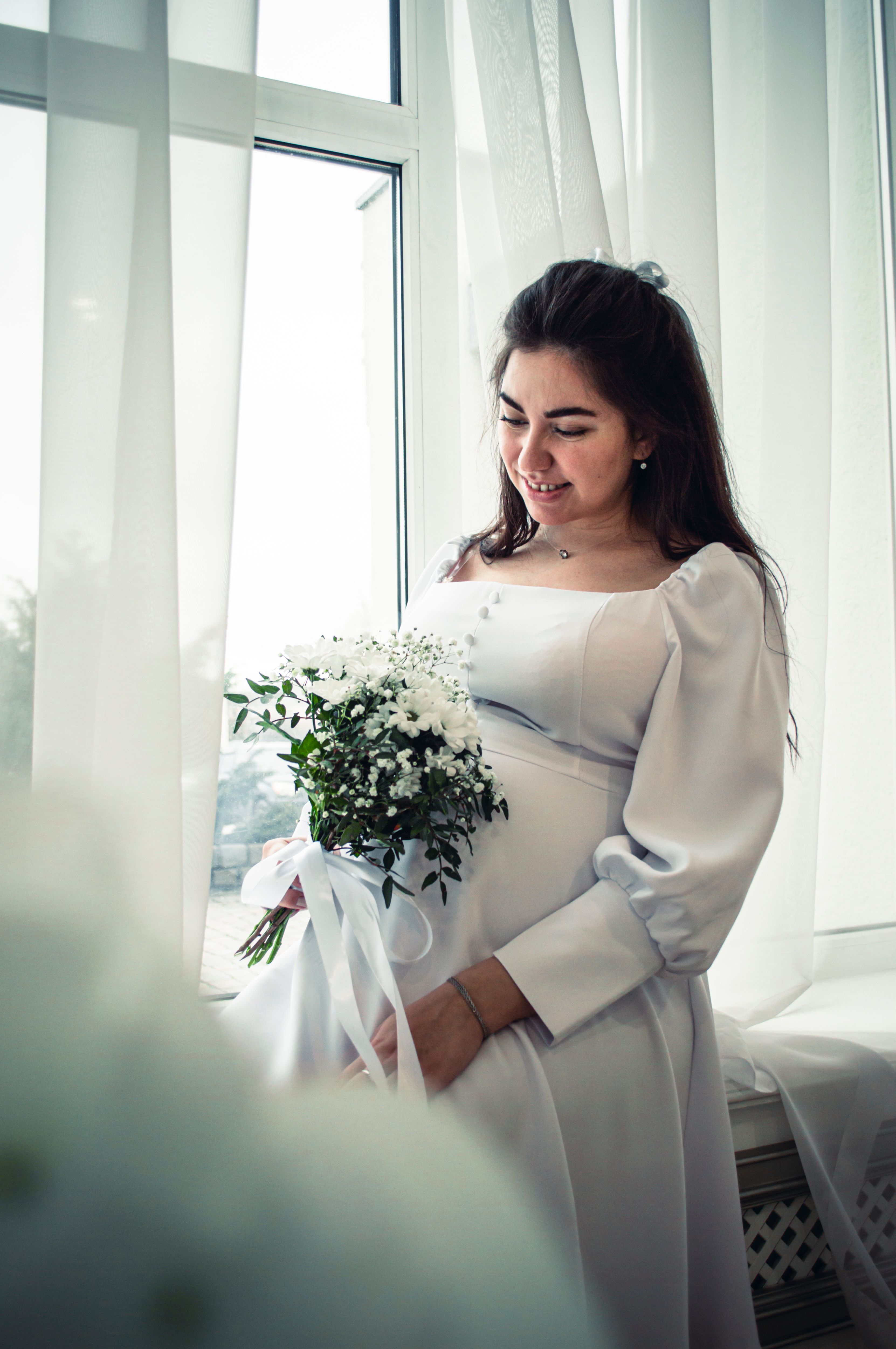 Elegant pregnant woman in white dress holding bouquet near window with white curtains.