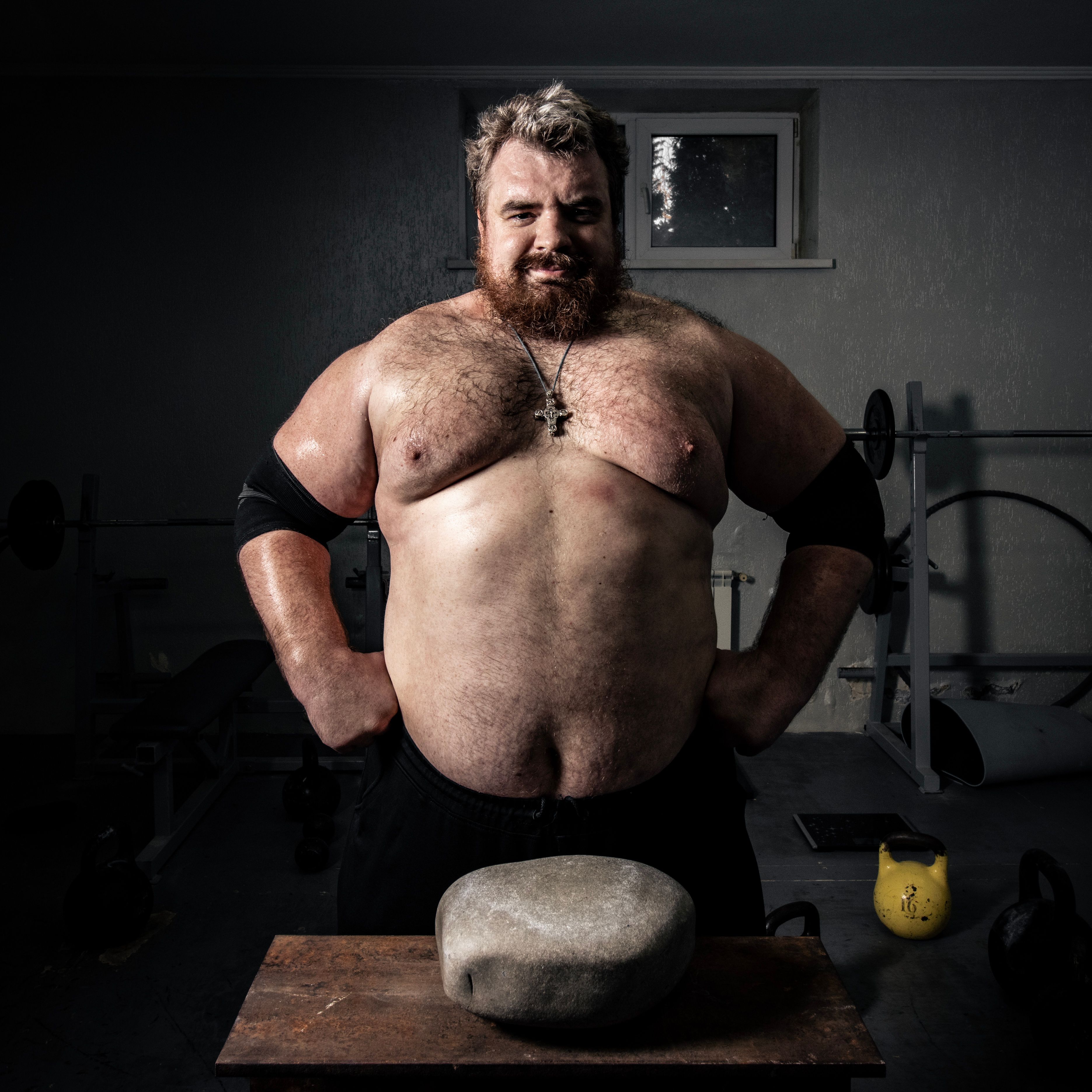 Bearded strongman posing with training stone in gym, embracing strength and power.