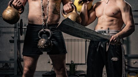 Two shirtless muscular men training in a gym with kettlebells and a sword, showcasing strength.
