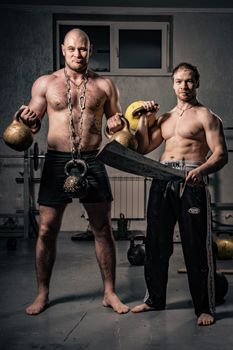 Two shirtless muscular men training in a gym with kettlebells and a sword, showcasing strength.