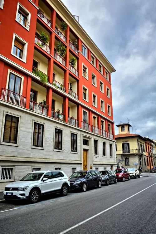 Free Urban city street scene featuring a vibrant red building with parked cars and cloudy skies. Stock Photo
