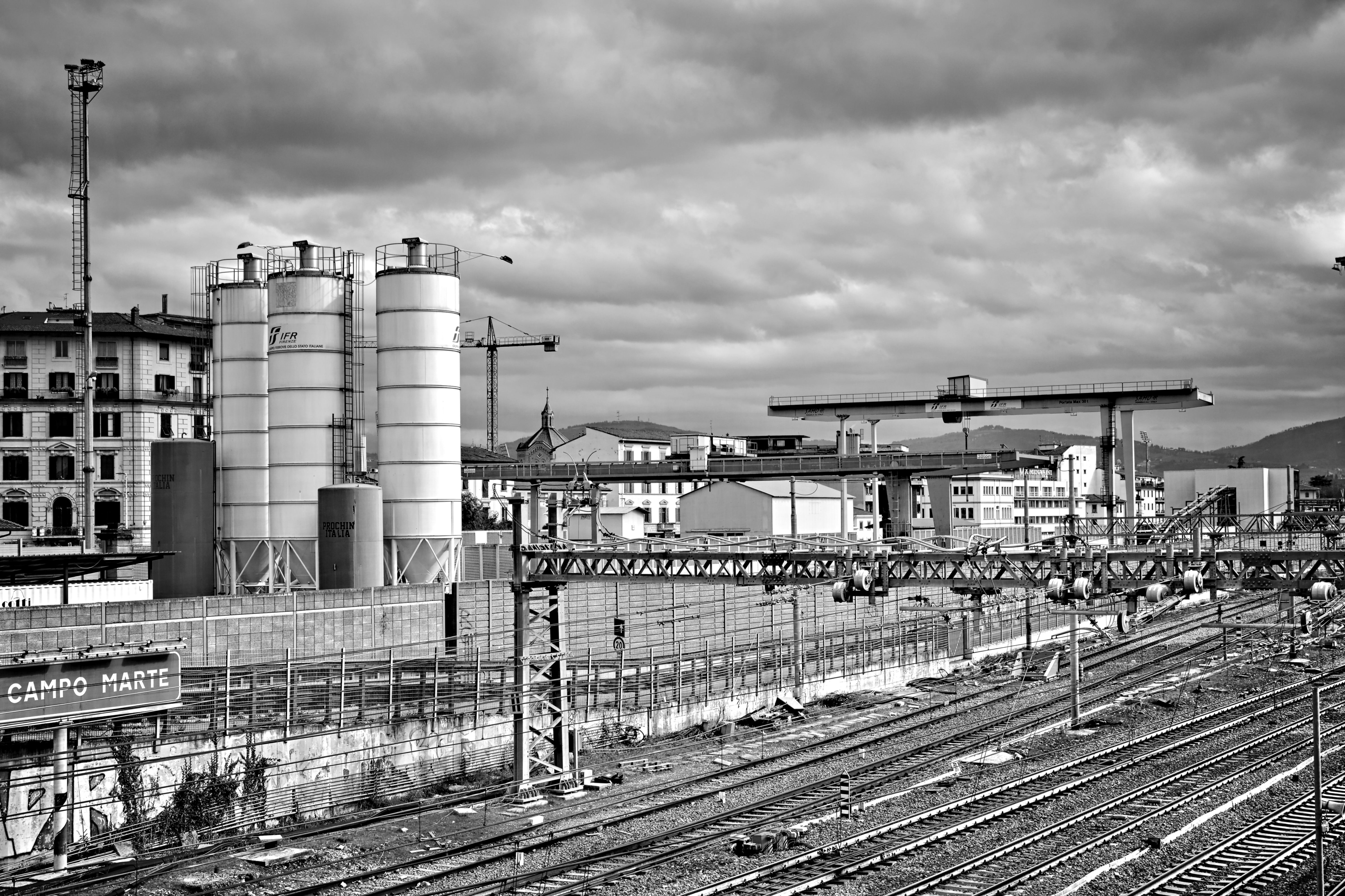 Black and white urban scene depicting a railway yard and industrial buildings under an overcast sky.