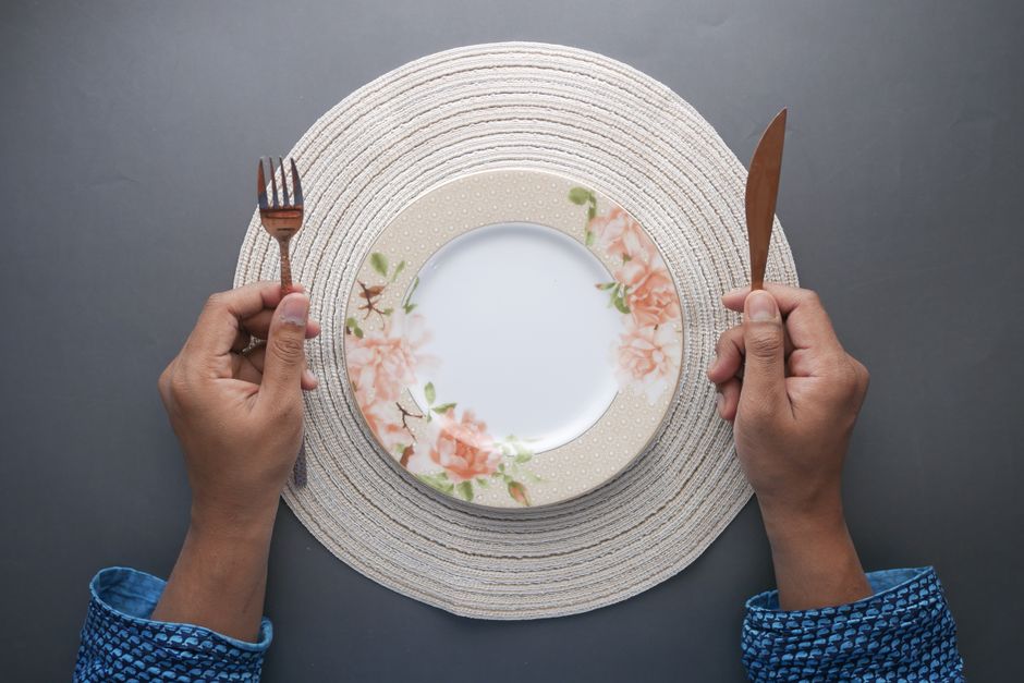Hands holding fork and knife around an empty floral plate on a woven placemat.