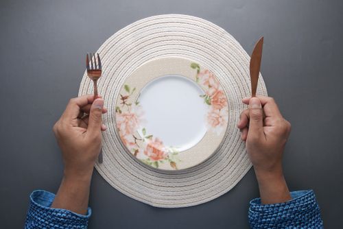 Free Hands holding fork and knife around an empty floral plate on a woven placemat. Stock Photo