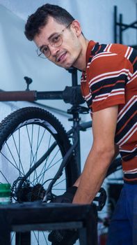 Smiling man wearing glasses repairing a bicycle indoors, showcasing a friendly and approachable demeanor.