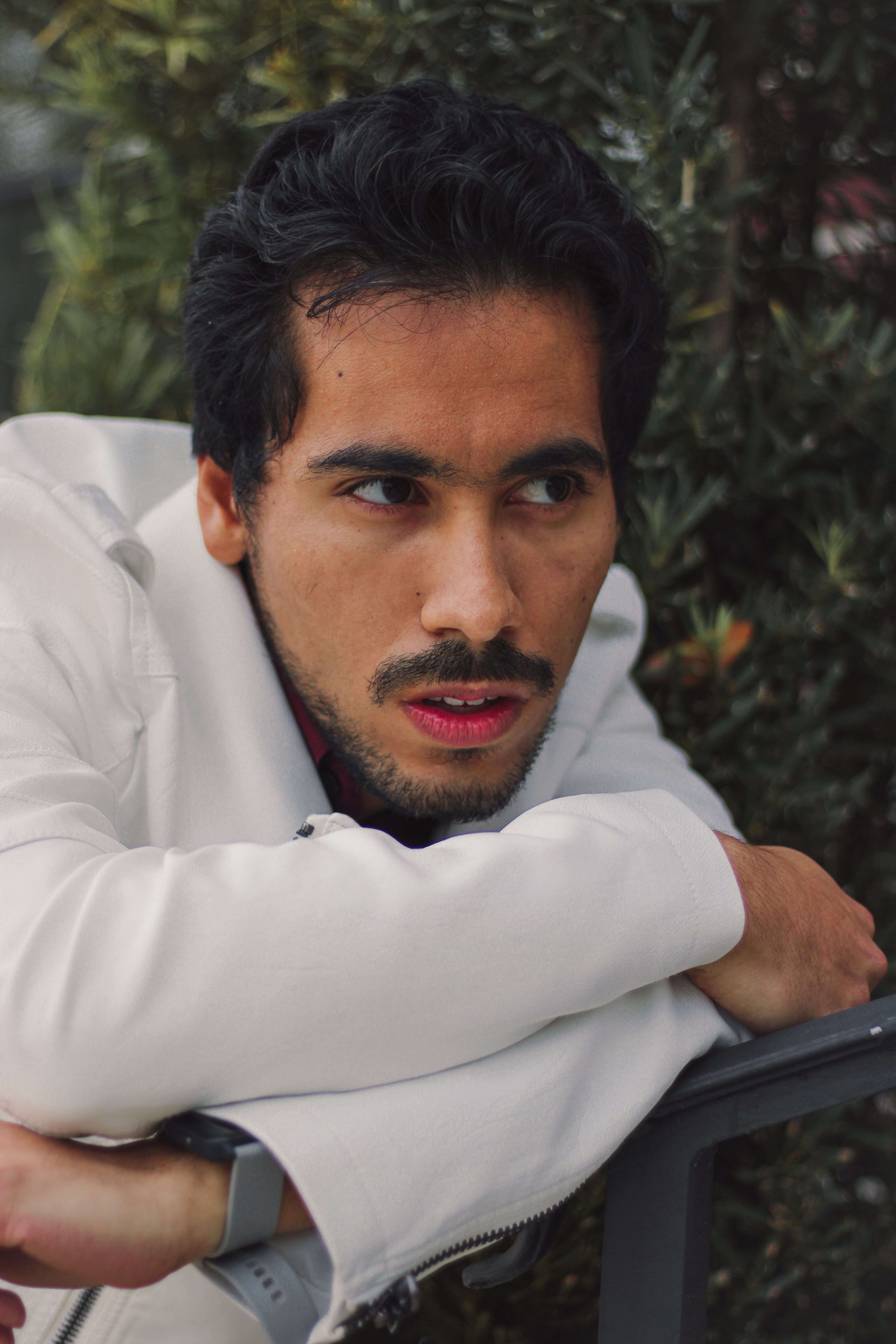 Pensive man leaning on railing amidst greenery, wearing white jacket.