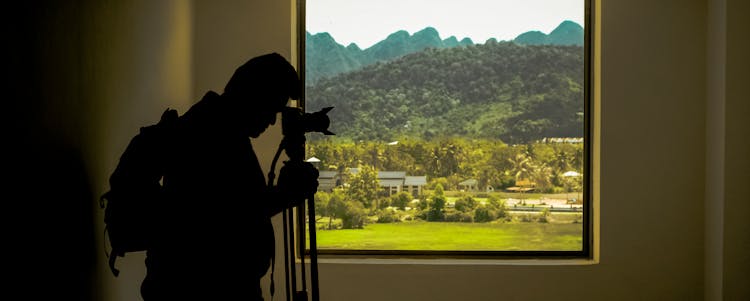 Silhouette Of Man Standing Near Window While Holding Camera With Tripod