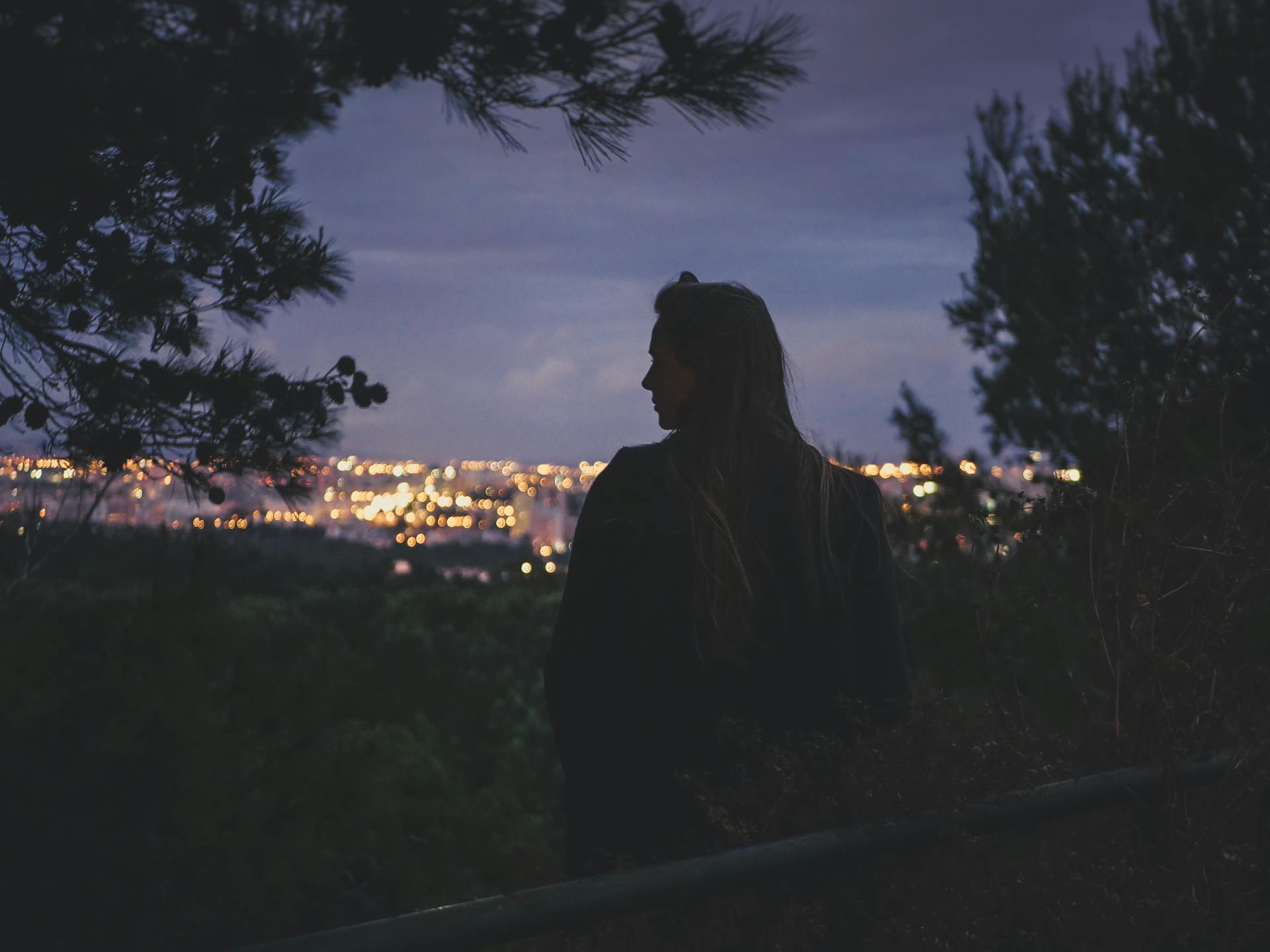 A woman gazes over the illuminated Lisbon cityscape at dusk, surrounded by trees.