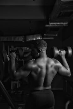 Black and white photo of a shirtless man lifting dumbbells in a gym, showcasing muscular physique.