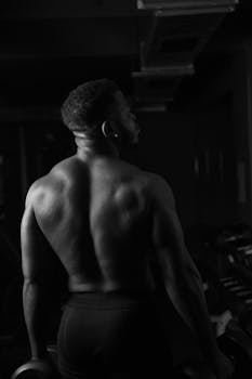 Black and white image of a muscular, shirtless man in a gym, showcasing strength and fitness.