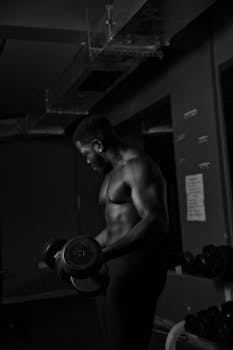 Monochrome photo of a shirtless bodybuilder exercising with dumbbells in a gym.