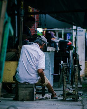 A senior craftsman sits on a stool working at an outdoor street workshop.