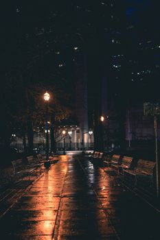 A moody night scene of a wet park path lined with benches and illuminated by streetlights.