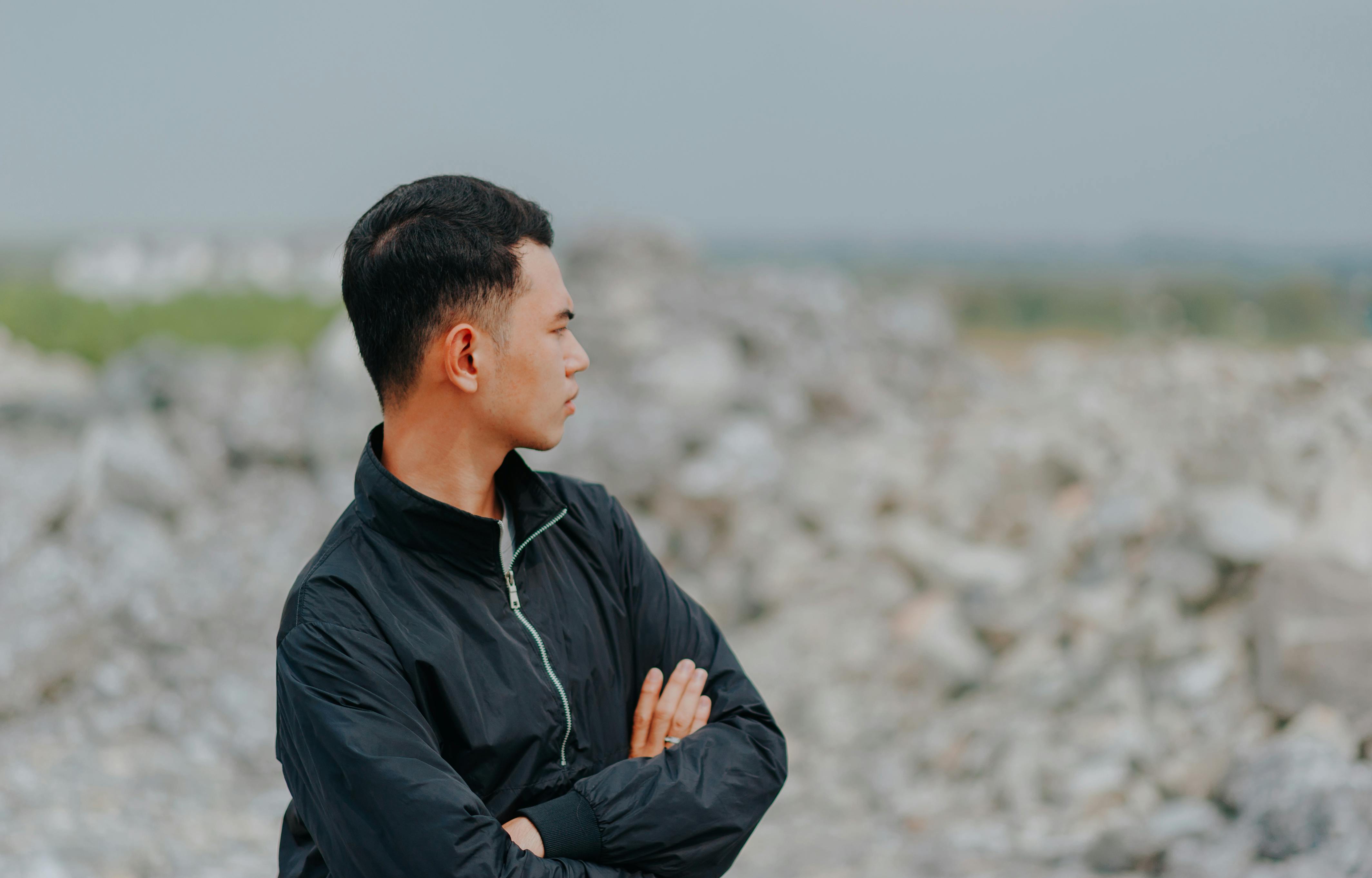 A man in a black jacket poses thoughtfully in a natural outdoor setting with a blurred background.