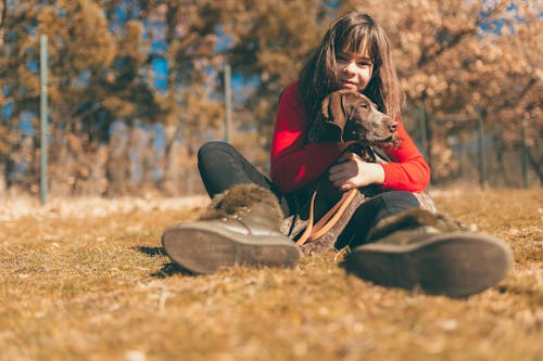 A Woman Sitting on Brown Grass Hugging a Brown Short Coated Dog