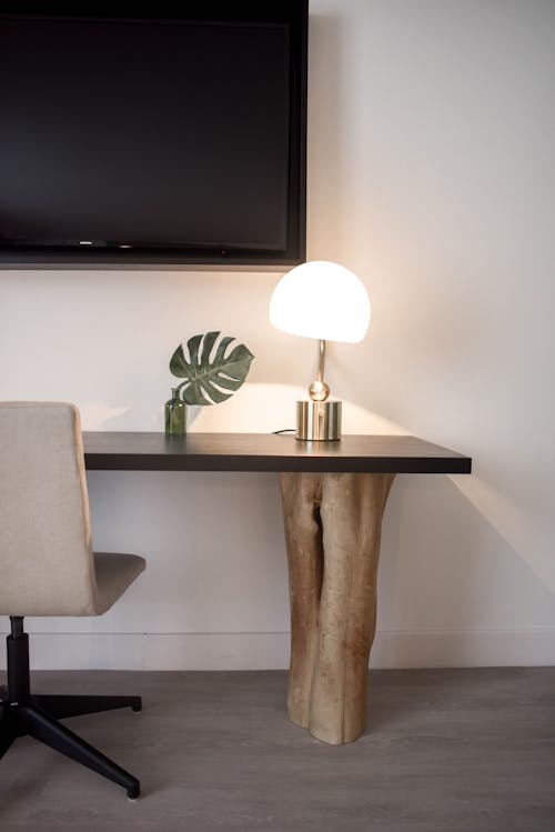 Lámpara De Mesa Con Pantalla Blanca Con Base De Acero Inoxidable En Un Escritorio De Madera Marrón Cerca De La Pared Pintada En Blanco Con Tv De Pantalla Plana Montada En La Pared