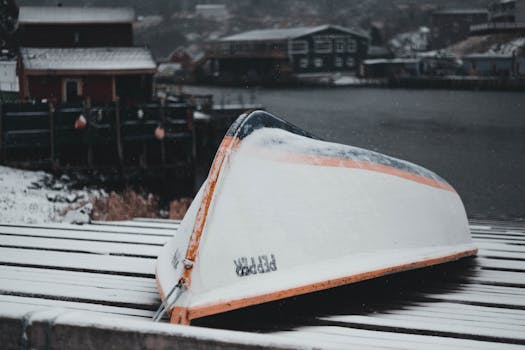 A snow-covered rowboat sits upside down by a snowy coastline in a quaint winter town.
