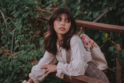 A young woman with bangs gazes thoughtfully in an outdoor garden setting.