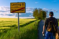 A Man Standing on the Paved Countryside Road Near the Farm Field and a Road Sign