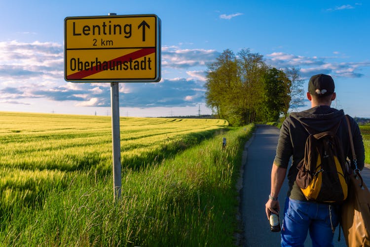 A Man Standing On The Paved Countryside Road Near The Farm Field And A Road Sign