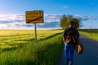 A Man Walking on the Paved Countryside Road Near the Farm Field and a Road Sign