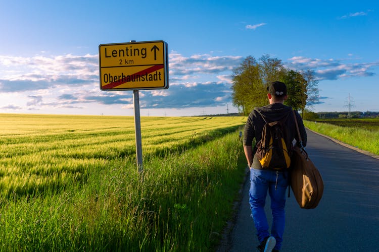 A Man Walking On The Paved Countryside Road Near The Farm Field And A Road Sign