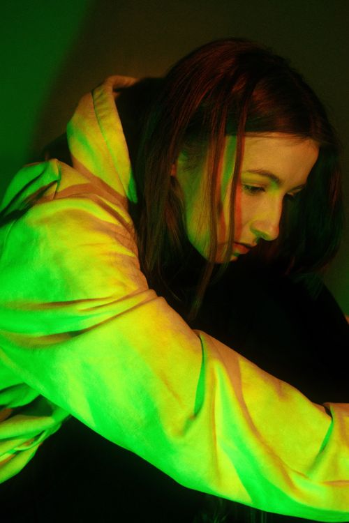 Free Pensive teenager in a green neon light, wearing a hoodie, deep in thought. Stock Photo