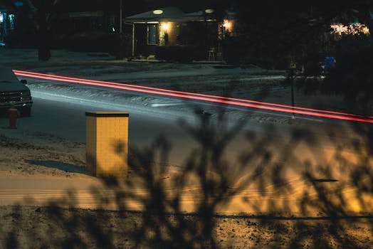 A snowy street at night captured with streaking car lights and parked cars under streetlights.