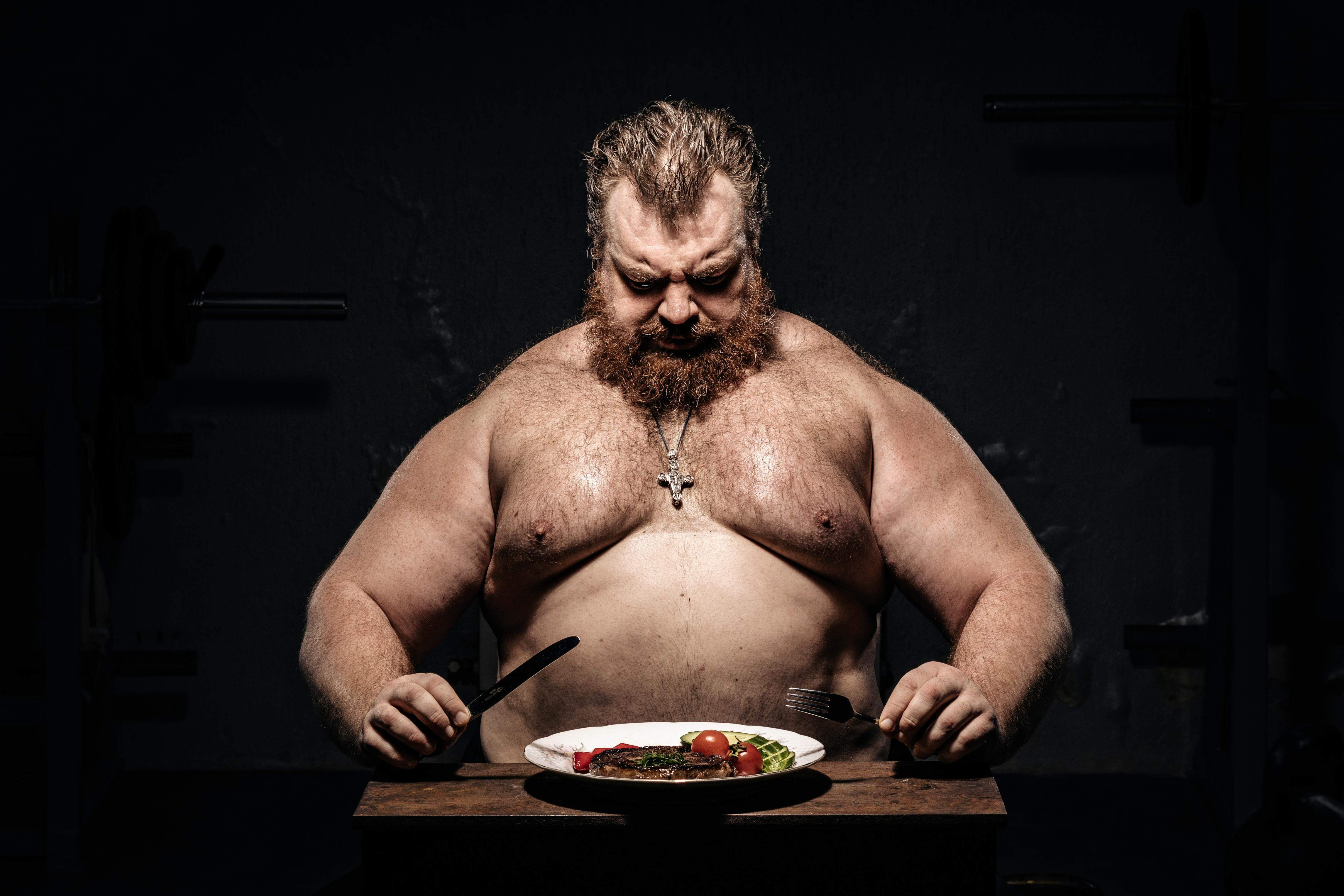 Bearded muscular man in dim light, preparing to eat steak with focus on fitness and nutrition.