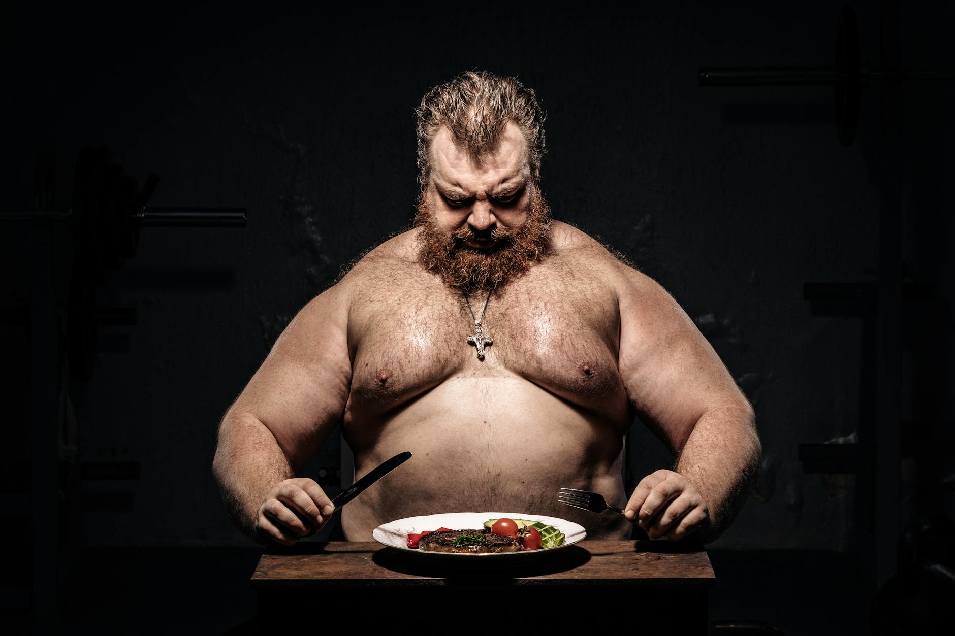 Bearded muscular man in dim light, preparing to eat steak with focus on fitness and nutrition.