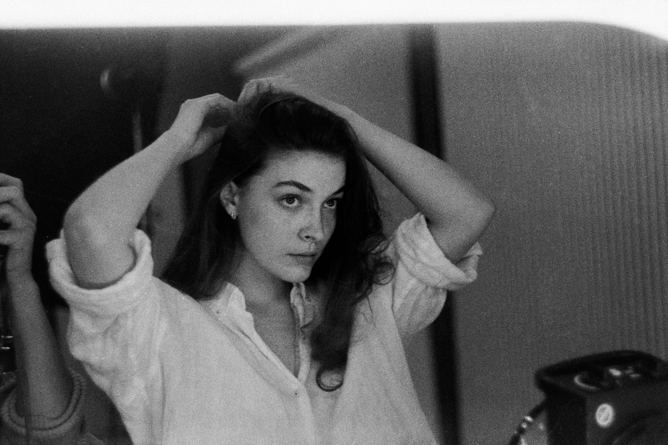 Black-and-white image of a woman in a white shirt adjusting her hair while looking in a mirror.