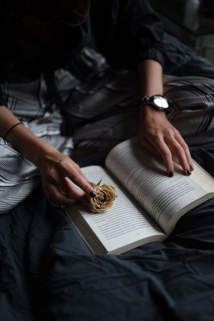 Hands holding a book with a dried flower on a dark cloth background.