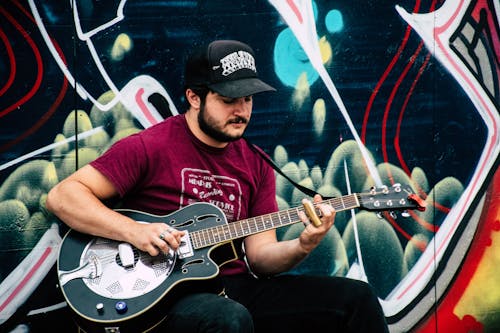 무료 검은 색과 녹색 그림이있는 벽 근처에서 공명기 기타를 연주하는 빨간색 셔츠를 입은 남자 스톡 사진
