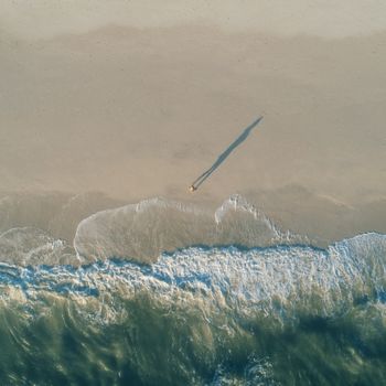Aerial view of a lone figure casting a long shadow on a serene beach with gentle waves.