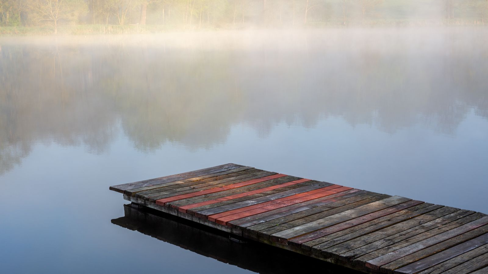 A wooden dock extending into a foggy lake surrounded by green trees, no people in the frame, a real stock photo that sets the scene for Crystal Lake, the Friday the 13th prequel set around a lake and a summer camp
