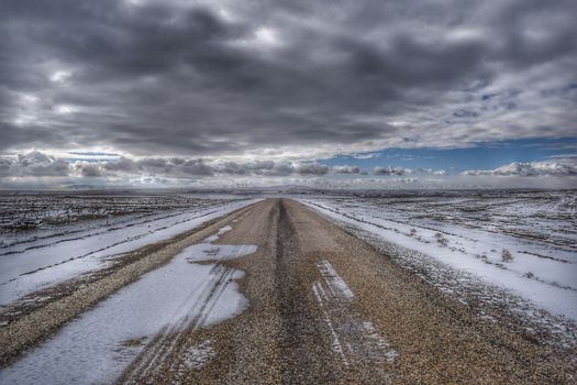 A deserted winter road stretches through snow-covered fields under a dramatic cloudy sky.