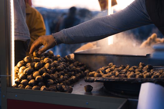A street vendor's hand reaches over a pile of roasted chestnuts at an outdoor market stall at night.
