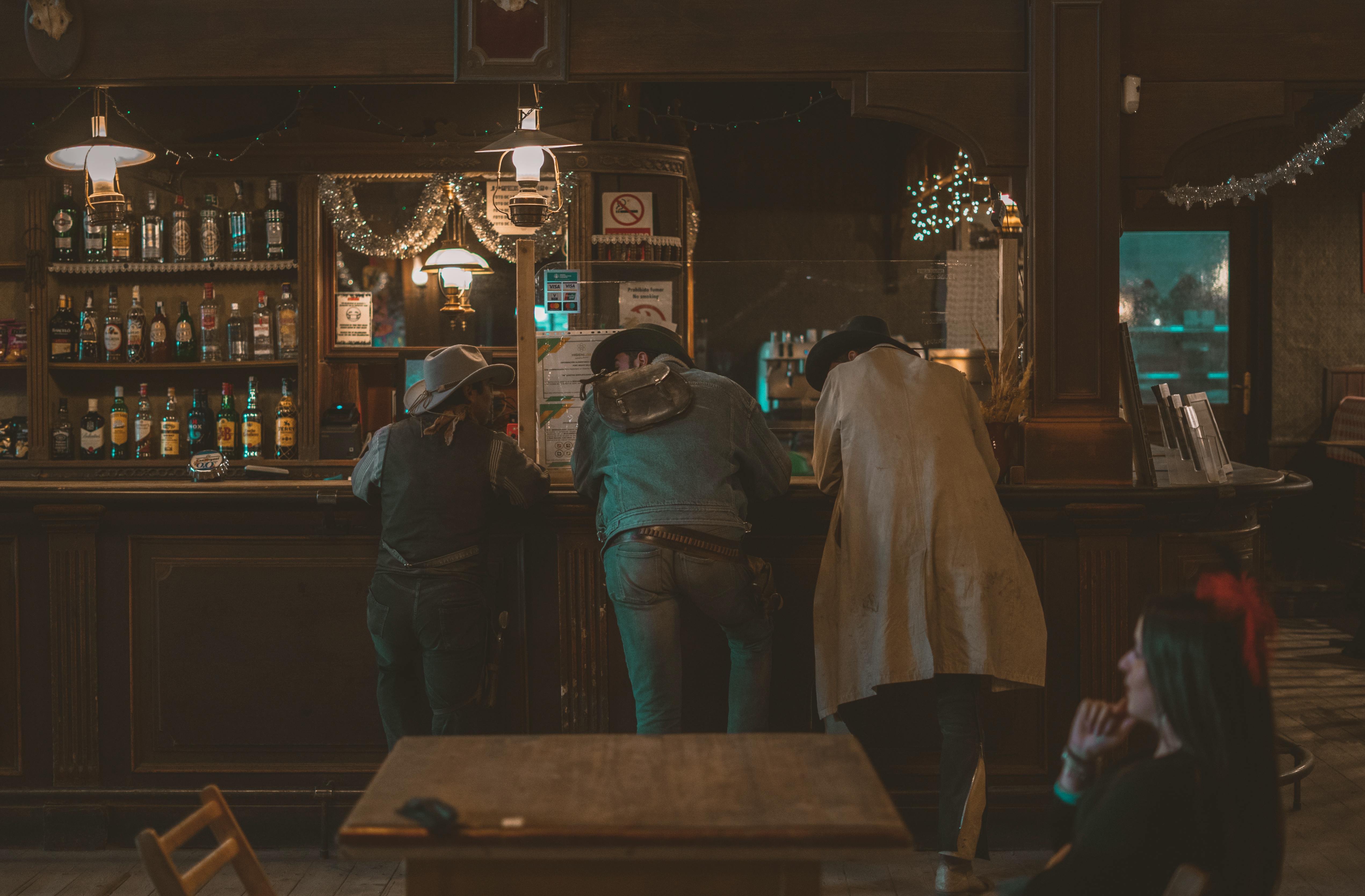 Three cowboys in hats stand at a rustic bar counter inside a western-themed saloon.