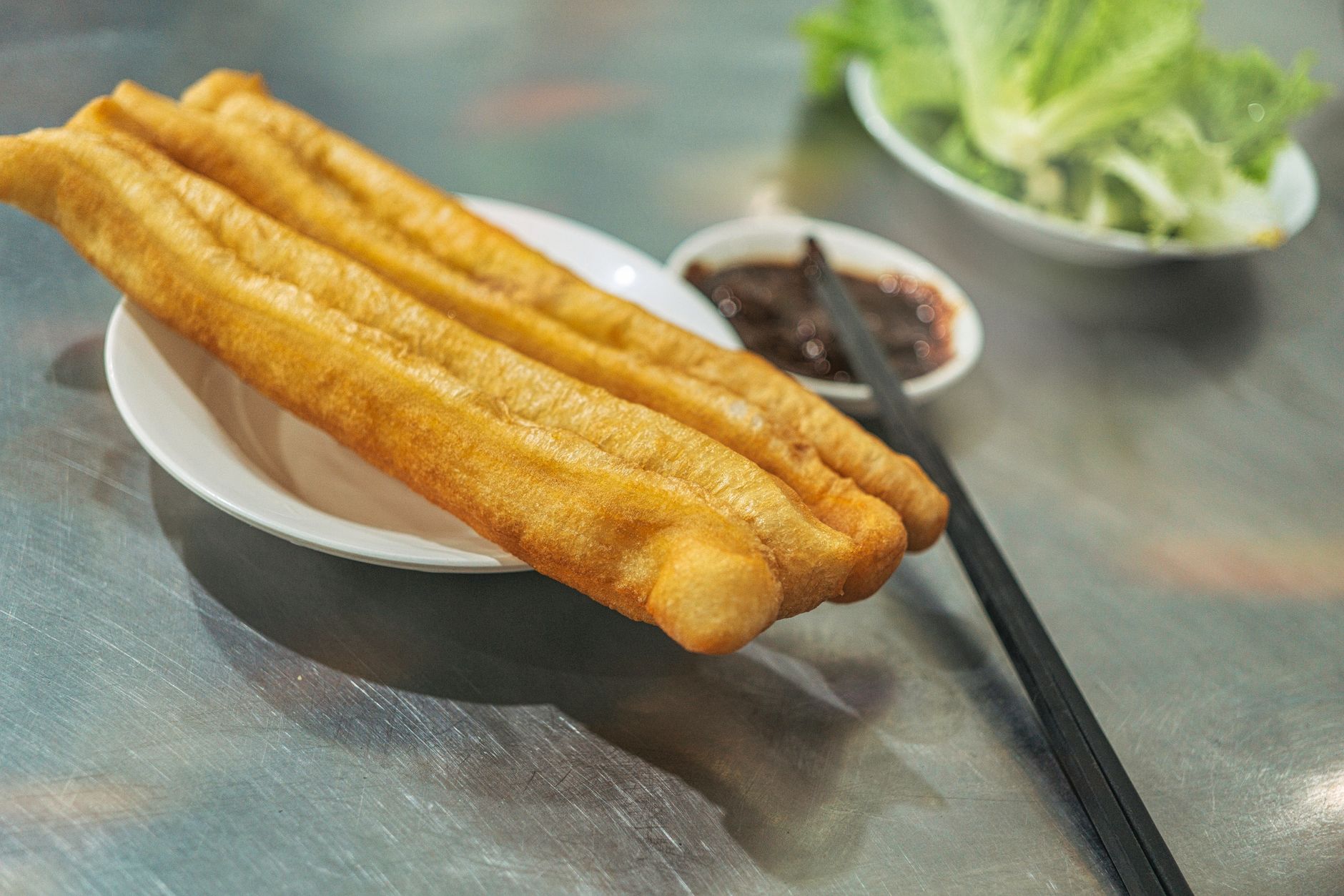 Close-up of crispy youtiao served with dipping sauce and chopsticks on a metal table.