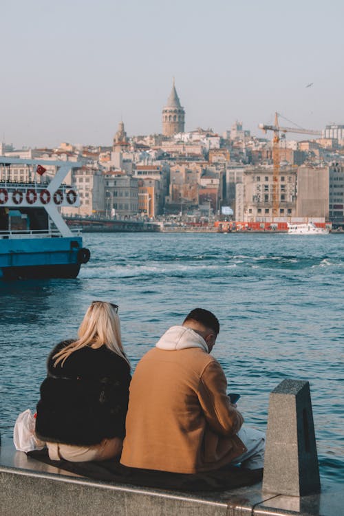 A Man and Woman Sitting Near the Body of Water with a City View