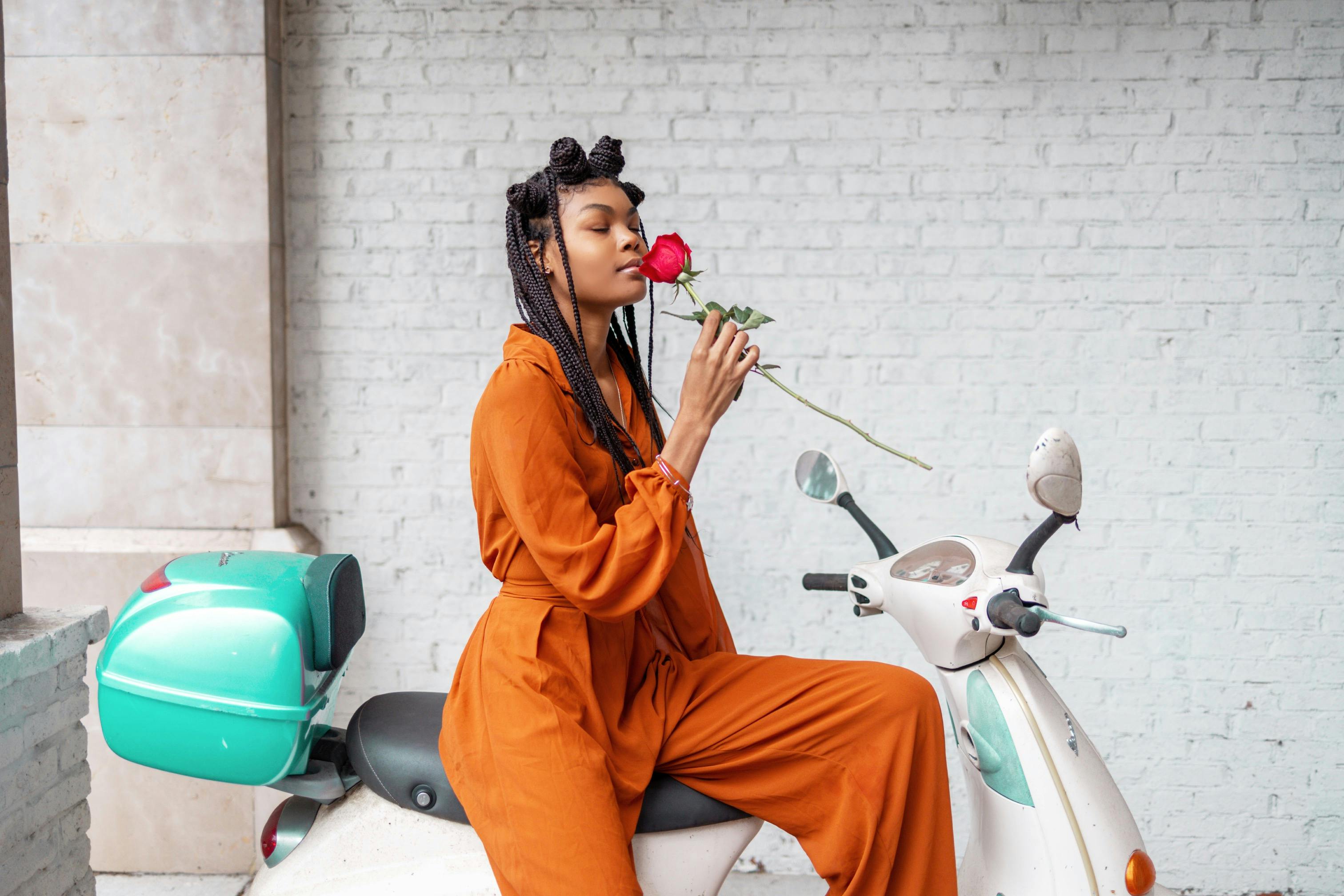 Young woman in an orange jumpsuit with braided hair enjoys the scent of a red rose while sitting on a scooter.