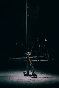 A lone electric scooter under a street lamp on a snowy night outside.
