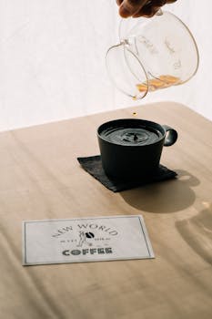 Hand pouring coffee into a mug on a sunny table with a New World Coffee card.