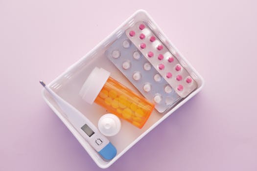 Overhead shot of medical supplies including pills and thermometer in a tray.