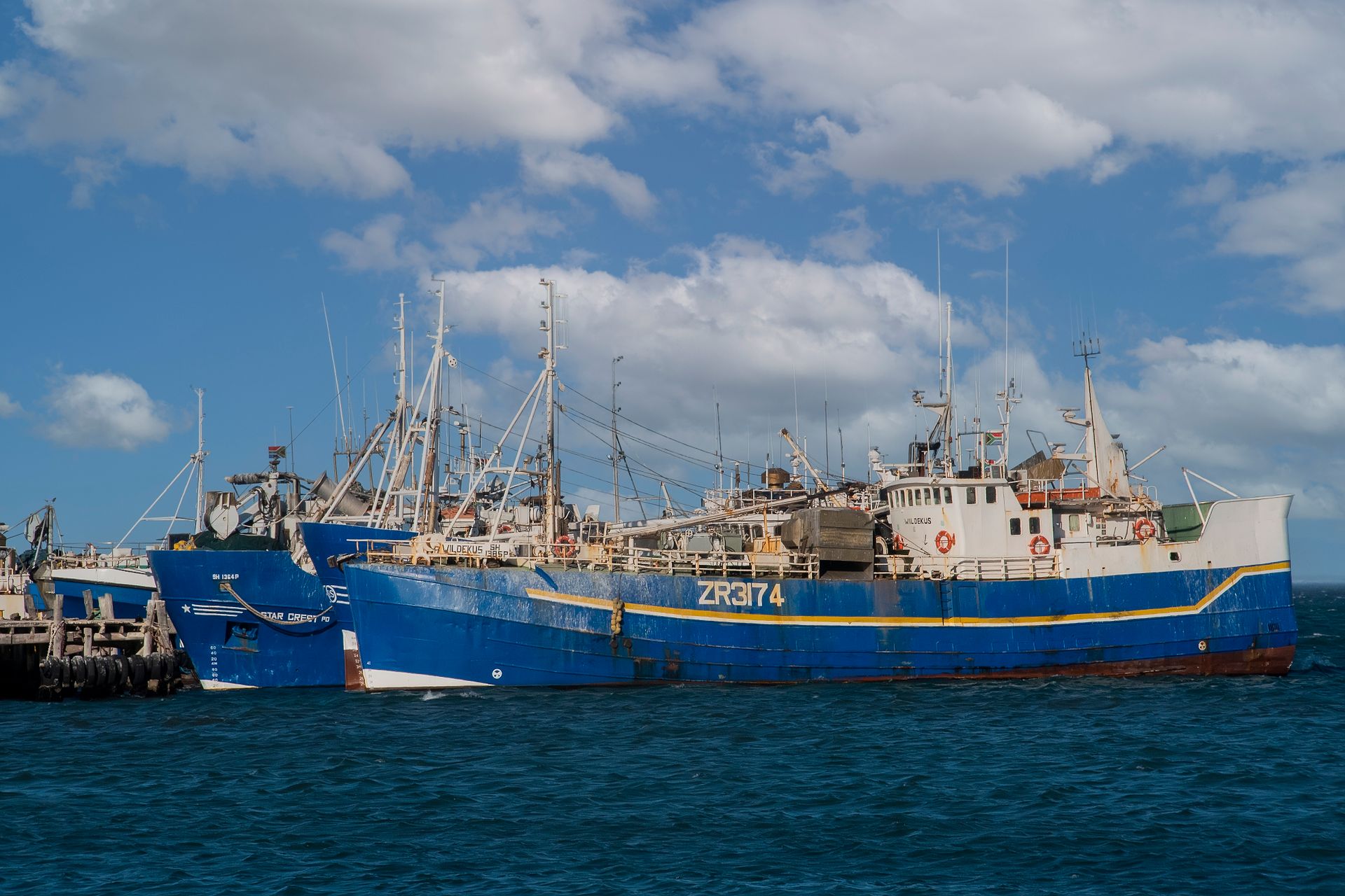 Fishing vessels with blue hulls docked at a pier under a cloudy sky.